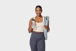 African American woman standing while holding a yoga mat in one hand and a water bottle in the other. She appears ready for a workout session or a yoga class, with a focused expression. photo