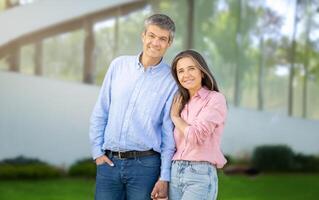 A joyful couple stands close together, sharing smiles in front of a striking modern building. The sunlight creates a warm atmosphere, highlighting their connection and happiness. photo