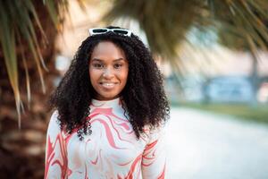 This image shows a woman with long, curly black hair smiling in front of palm trees. She is wearing a white and pink shirt with a marbled pattern and has a pair of sunglasses on top of her head. photo