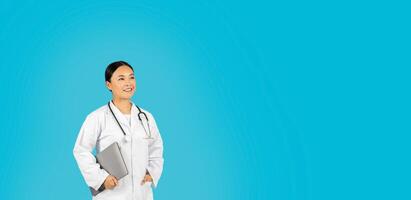 Asian woman doctor, wearing a white coat and a stethoscope, smiles while holding a laptop in her left hand and looking at copy space. She is standing in front of a light blue background. photo