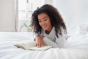 A Hispanic woman is laying comfortably on a bed, engrossed in reading a book. The room is softly lit, and she is fully focused on the text, turning pages occasionally. photo
