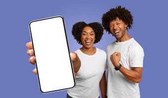 A happy African American couple stands in front of a purple background, with the woman holding a smartphone with a blank screen towards the camera and the man raising a fist in celebration photo