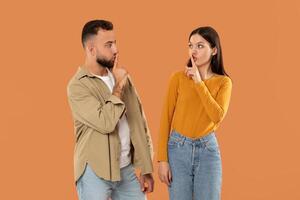 A young man and woman stand facing each other, both holding a finger to their lips, signaling for silence against a solid orange background. They seem to be engaged in a quiet conversation or secret. photo