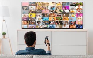 A man sits on a couch in his living room, facing a large television displaying many different thumbnails on screen. He is holding a television remote in his hand, selecting something to watch photo