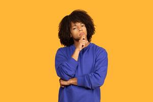 This image features a young man with curly black hair wearing a blue sweater. He is standing in front of a bright yellow background and looking intently at something off-camera. photo