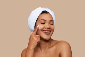A woman with a towel wrapped around her head is gently applying cream to her face. She is standing in front of a mirror, focusing on her skincare routine. photo