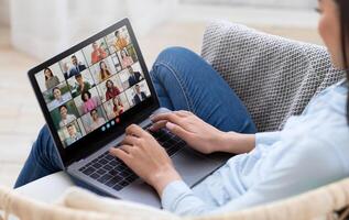 A woman is sitting comfortably on a couch, using her laptop to join a virtual team meeting. The laptop screen shows a multi-person conference with various participants in different locations. photo