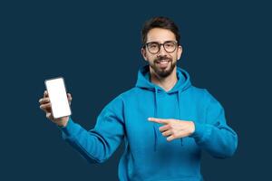 A cheerful young man with a beard and glasses is wearing a blue hoodie and presenting a smartphone with its screen facing forward towards the viewer. He is pointing at the screen photo