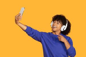 A young man with curly hair is taking a selfie on a yellow background. He is wearing headphones and smiling happily. His index finger is pointed towards the camera as if he is giving a thumbs up. photo