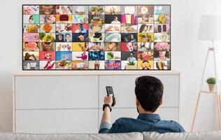 A man sits on a couch in a living room, facing a large flat screen television. He is holding a remote control in his right hand. The television screen displays a grid of colorful, diverse images. photo