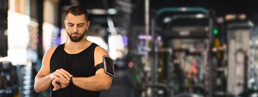 A young man with a beard is wearing a black tank top and checking his smartwatch. He is standing in a gym, with exercise equipment blurred in the background, copy space photo