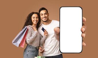 A happy couple holding shopping bags and a smartphone with a blank white screen. The man is holding the phone in front of him with his right hand, while the woman is holding a credit card and smiling photo