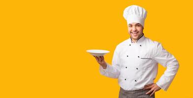 Bon Appetit. Smiling Cook Guy Holding Empty Plate Looking At Camera Standing Over Yellow Studio Background. Panorama photo