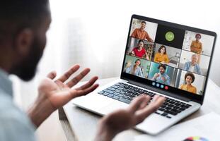 Black man sits in front of a laptop, participating in a conference meeting with several colleagues. He has his hands raised as if he is making a point during the call. photo