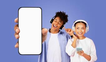 A young multiracial couple wearing headphones and smiling happily while listening to music on their smartphones, with a blank smartphone screen prominently displayed in the foreground. photo