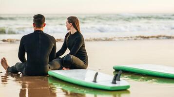 A couple in wetsuits are sitting on a beach with their surfboards. They looking at each other and appear to be in conversation. The beach is sandy and there is gentle wave breaking in the background photo