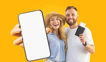 A young couple stands together against a bright yellow background. The woman is smiling broadly and holding a smartphone with a blank screen while the man is holding a passport and boarding pass. photo