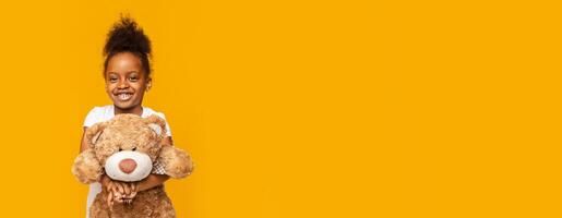 A joyful young African American girl stands confidently while hugging a large plush bear. She has a big smile on her face, radiating happiness. The background is vivid yellow, copy space photo