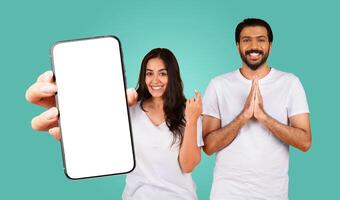 Eastern couple stand in front of a teal background. The woman is holding a smartphone with a blank white screen, pointing at it with her left hand. The man stands behind her with his hands clasped photo