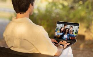 A man sits on a bench in a park and chats with a group of friends on his laptop. The man is facing away from the camera, and the screen of the laptop shows the friends smiling and waving photo