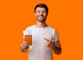 A smiling man in a white t-shirt holds a passport and airplane tickets in his left hand and points to the right with his right hand, against a bright orange background. photo