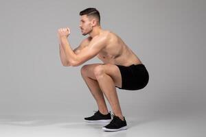 A man is seen on a grey background doing a squat exercise, bending his knees and lowering his hips while keeping his back straight. He is engaged in a strength training routine for lower body muscles. photo