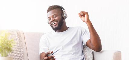A happy Black man is sitting on a couch and listening to music on his smartphone through headphones. He is smiling and bobbing his head to the music. photo