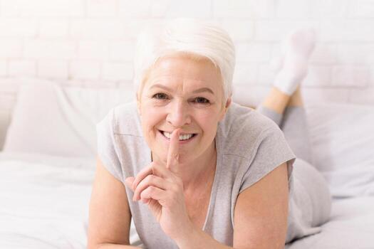 un alegre mayor mujer con corto blanco pelo mentiras en su estómago en un cama, su ojos radiante con alegría. ella lugares su dedo en su labios, sugerencia un juguetón secreto o un tranquilo momento foto