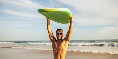 A man with a bare chest and sunglasses is standing on a sandy beach with blue water and white waves behind him. He is holding a green surfboard above his head with both hands, smiling at the camera. photo