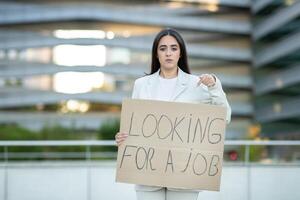 A woman wearing a white blazer stands in front of a modern city building, holding a cardboard sign that reads Looking For A Job. She points at the sign with her right hand photo
