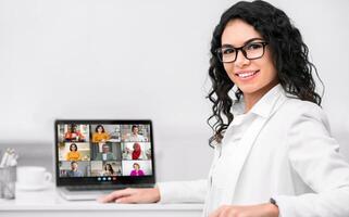 A woman in a white blazer sits at a desk in front of a laptop computer and smiles at the camera. The laptop screen displays a conference with multiple participants photo