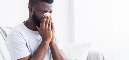African American man sits on a bed, his head bowed, blowing his nose into a tissue. He appears to be suffering from a cold or allergies. The room is well-lit and has a white background, copy space photo