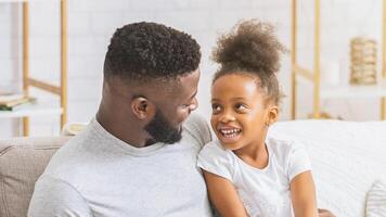 A close-up image of a happy Black father and daughter laughing together on a couch in a living room. The two are enjoying each others company and are clearly having a good time. photo