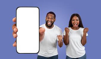 Black couple stand in front of a purple background, holding a smartphone with a blank screen. The man is pointing at the phone with his left index finger and has a huge smile on his face. photo