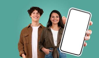 Multiracial couple stand against a teal backdrop. The woman holds a large smartphone with a blank screen in her right hand, extending it out towards the viewer. Both are smiling. photo