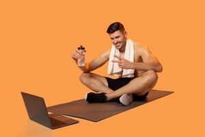 A young man is sitting cross-legged on a yoga mat, engaging in a virtual workout session. He wears a towel around his neck and holds a water bottle, smiling and gesturing towards his open laptop. photo