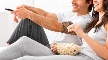 A young couple is relaxing on a couch, watching television and enjoying a bowl of popcorn. The man is holding a remote control, while the woman is reaching for a handful of popcorn, cropped photo