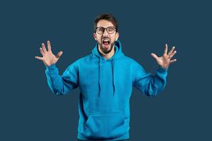 un Encantado joven hombre con lentes es en pie en contra un oscuro azul fondo, su boca amplio abierto como Si gritos con alegría o sorpresa. él es vistiendo un brillante azul capucha y tiene elevado su manos foto