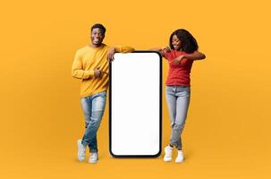 A cheerful black couple stands to the left and right of a large smartphone mockup with a blank white screen, set against a vibrant yellow background. The man points at the screen photo