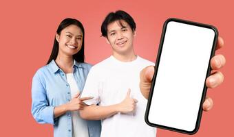 Two young Asian people stand in front of a red background. The man is holding a smartphone with a blank screen in his right hand, and he is pointing at the screen with his left hand. photo