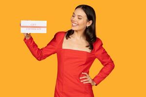 This image shows a woman wearing a red dress standing against a yellow background. She is holding a white gift certificate in her right hand, and looking at it with a smile. photo