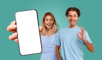 A couple poses happily in front of a teal background. The woman holds a smartphone with a blank screen and is smiling excitedly, while the man gives a thumbs up, showing his approval. photo