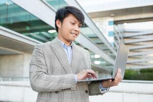 A young Asian man in a plaid suit stands outside a modern building, working on his laptop. The man is smiling and focused on his work, and the building in the background is a sleek, modern structure photo