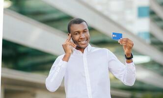 Black man in white shirt stands outside and smiles while holding a credit card in his right hand and talking on his cell phone. He is looking at the camera and standing in front of a modern building. photo