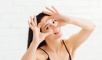 A cheerful woman wearing a black dress is making a heart shape with her hands while smiling. She is indoors, positioned against a bright, light-colored wall, and seems to be enjoying the moment. photo