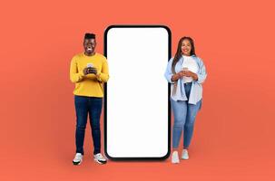With joyful faces, the young black man and woman pose beside a blank-screened, life-sized smartphone, emphasizing the endless possibilities for customization. photo