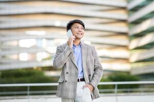 A young Asian man dressed in a grey plaid blazer and white pants is standing outside a modern building. He is smiling while talking on a white smartphone. photo