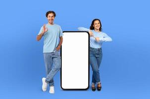 Smiling man and woman are standing side by side. They are looking at a blank screen of a phone held by the woman. The man appears to be pointing or gesturing towards the screen. photo