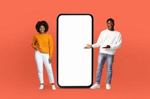 On a deep red background, a black lovers, both smiling, stands adjacent to an enormous smartphone with a blank display, ready for showcasing apps or designs. photo