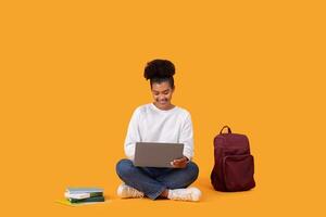 A young woman is sitting on a yellow background, using a laptop computer. A backpack is sitting on the floor next to her, and a stack of books is in front of her. photo
